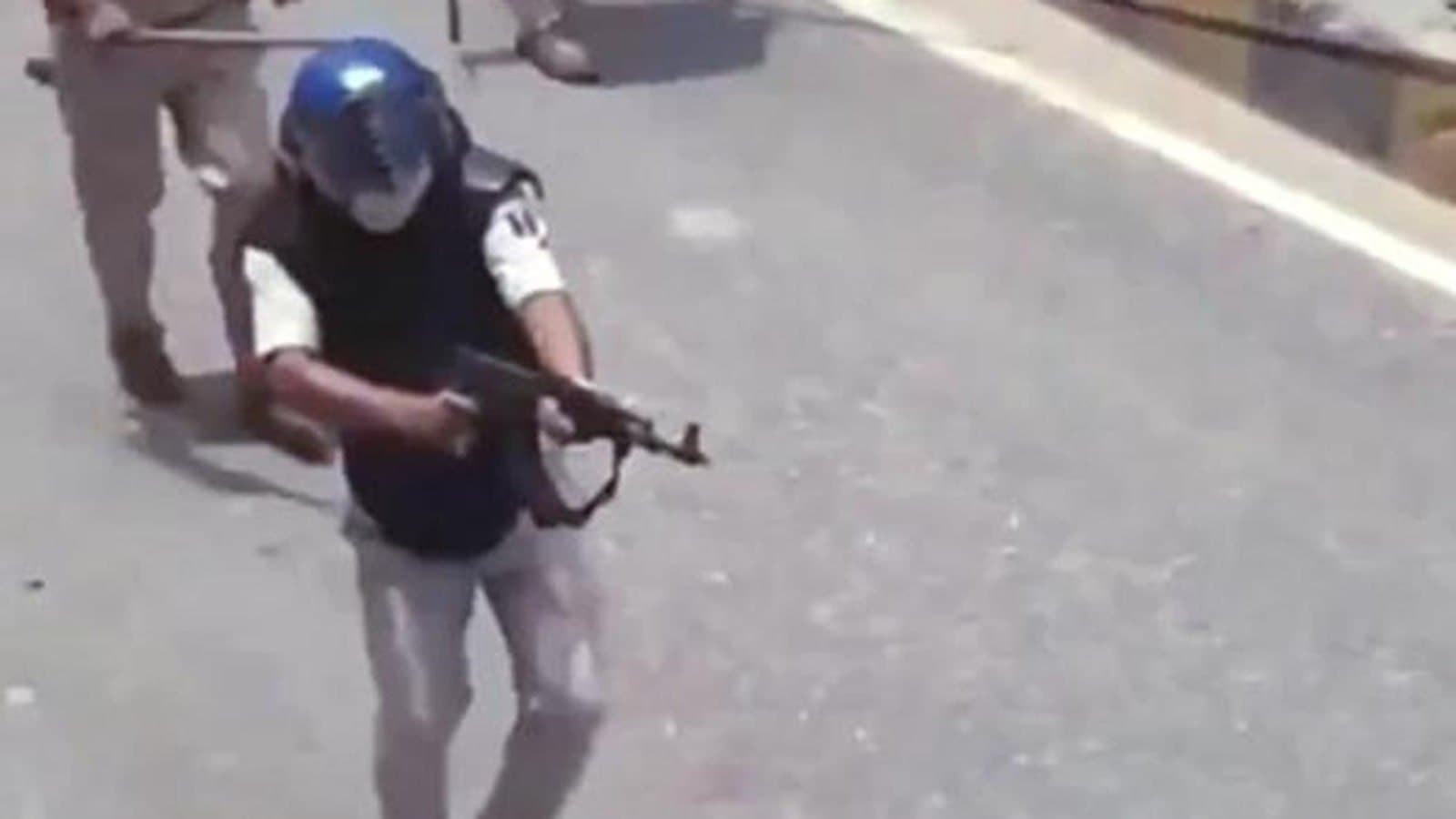 A video still of a policeman in a helmet and body armour crouching on a road holding an assault rifle, with another officer holding a lathi behind him.