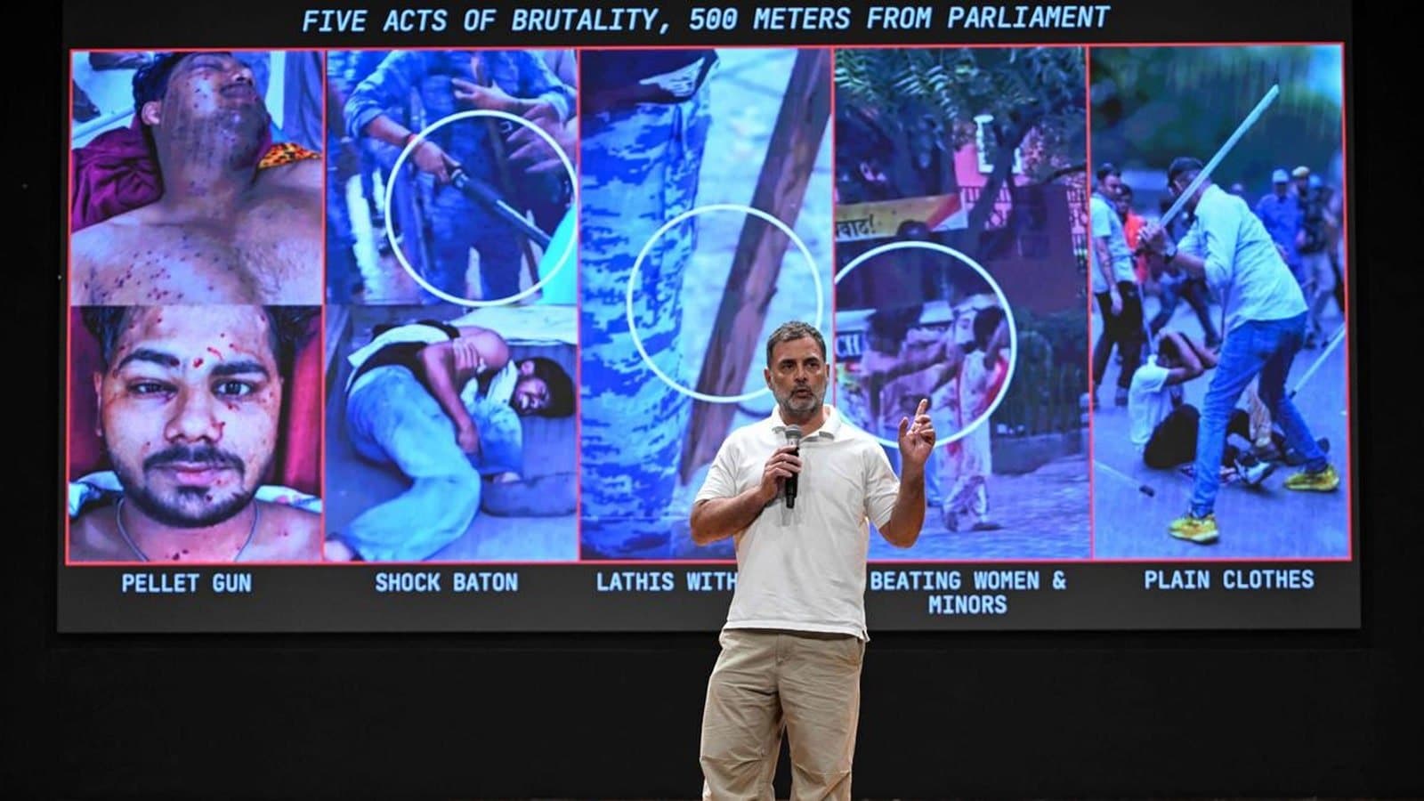 Rahul Gandhi speaking on stage with a microphone in front of a large screen displaying photographs from the protests under the heading Five Acts of Brutality, 500 Meters From Parliament.