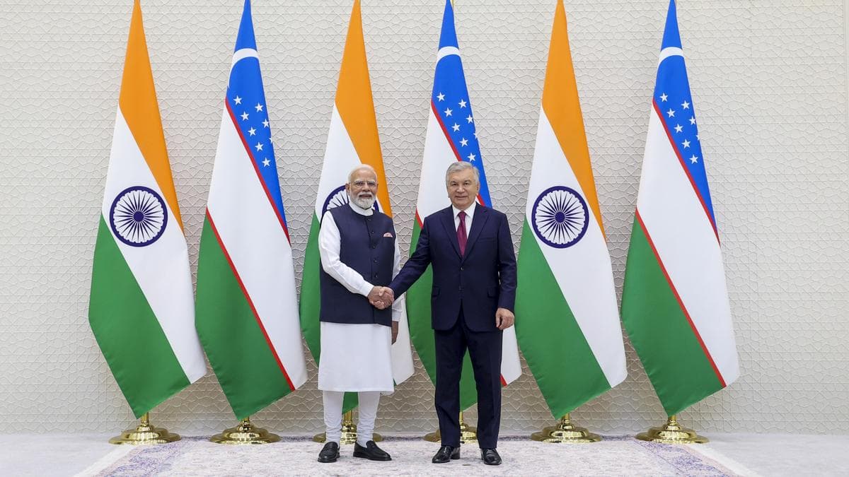 प्रधानमंत्री नरेंद्र मोदी भारतीय और उज्बेक झंडों के सामने उज्बेकिस्तान के राष्ट्रपति शवकत मिर्जियोयेव से हाथ मिलाते हुए